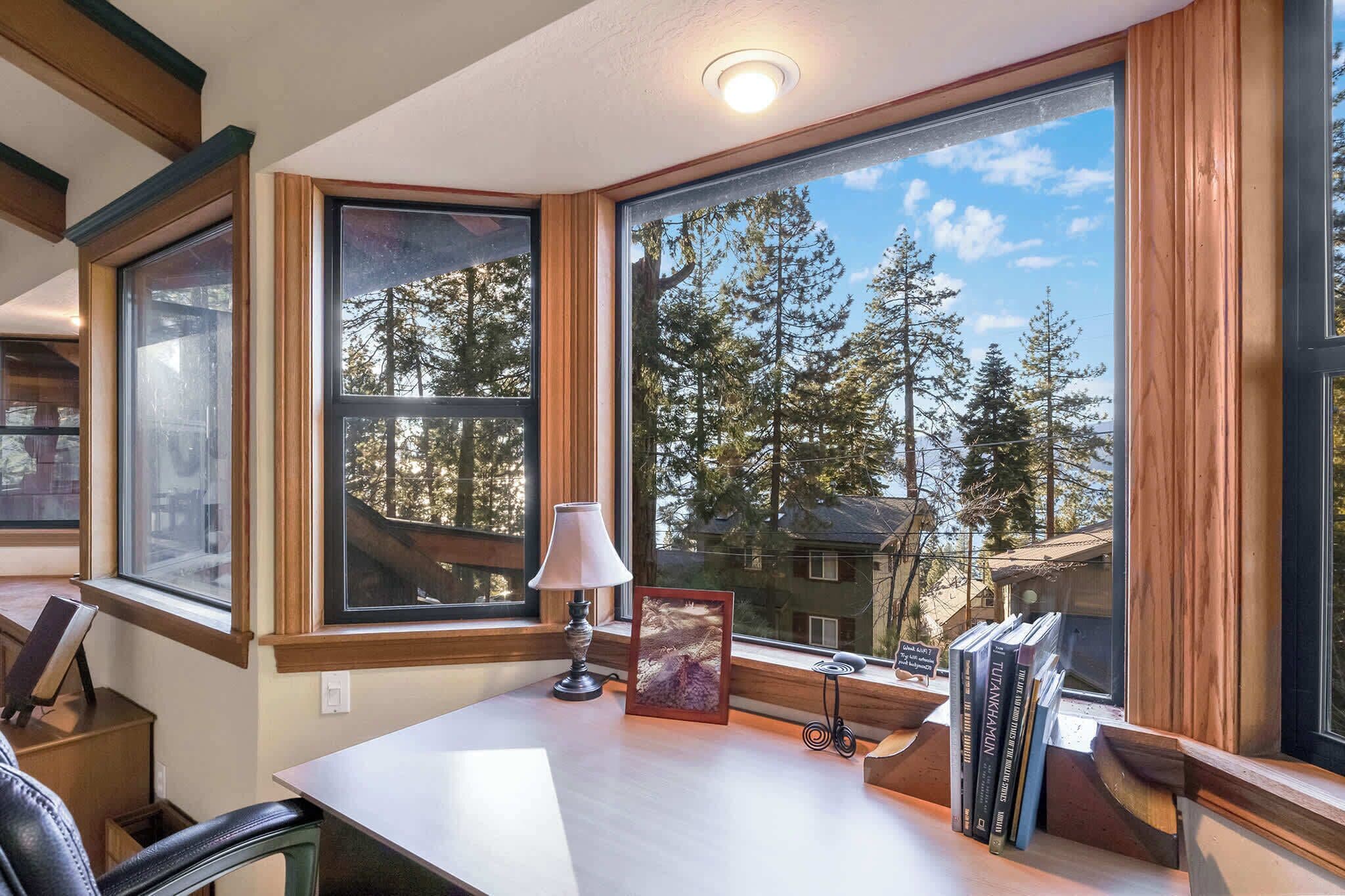 A room with large windows, a desk, a lamp, a framed picture, and books.