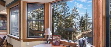 A room with large windows, a desk, a lamp, a framed picture, and books.