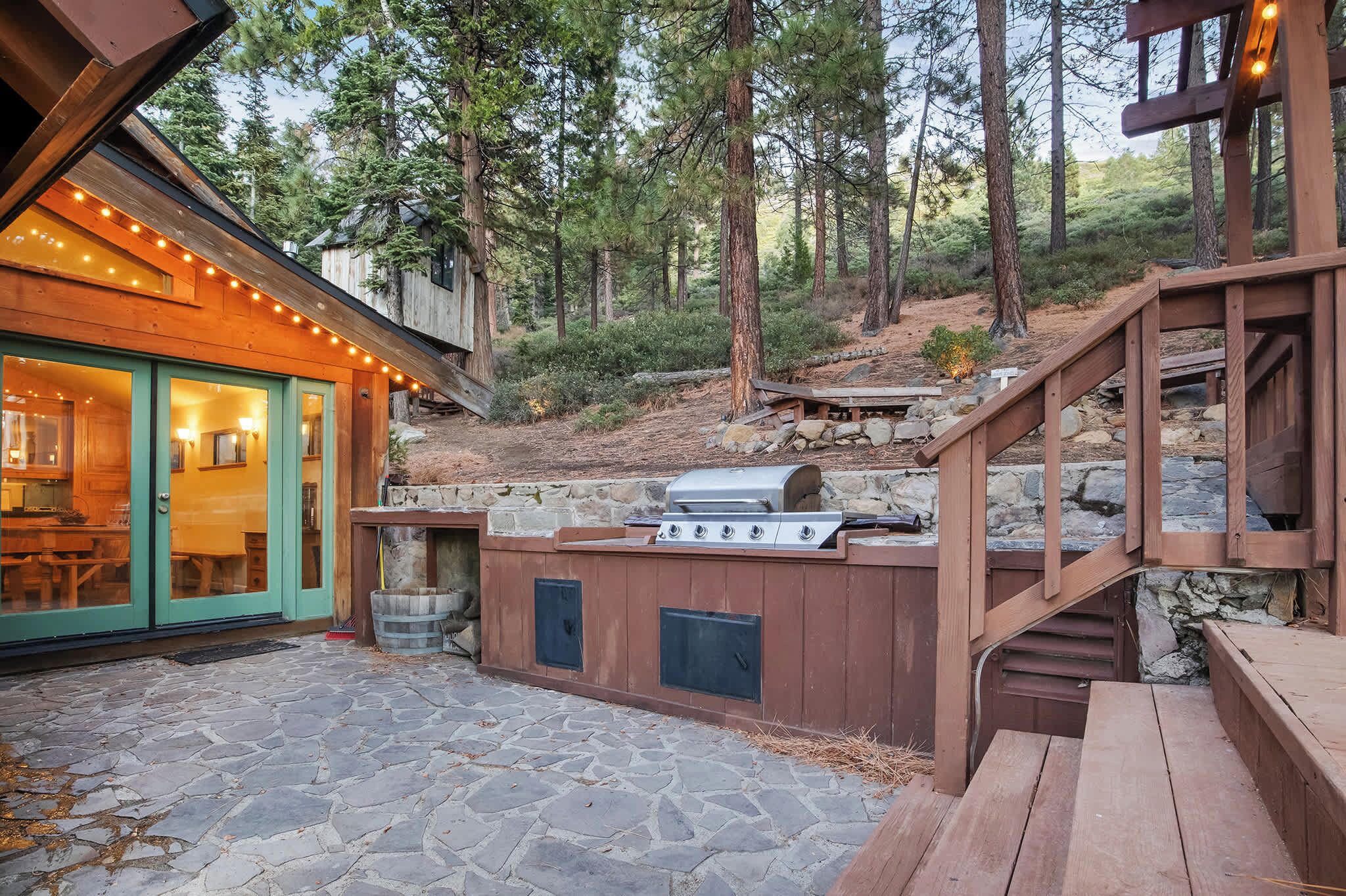 A wooden deck with a stone patio, a barbecue grill, and a wooden staircase leading to a deck area.