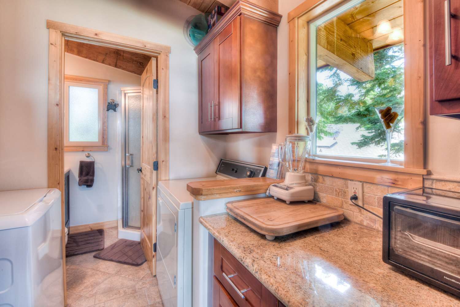 A compact kitchen with wooden cabinets, a countertop with a blender, a window with a view, and a door leading to another room.