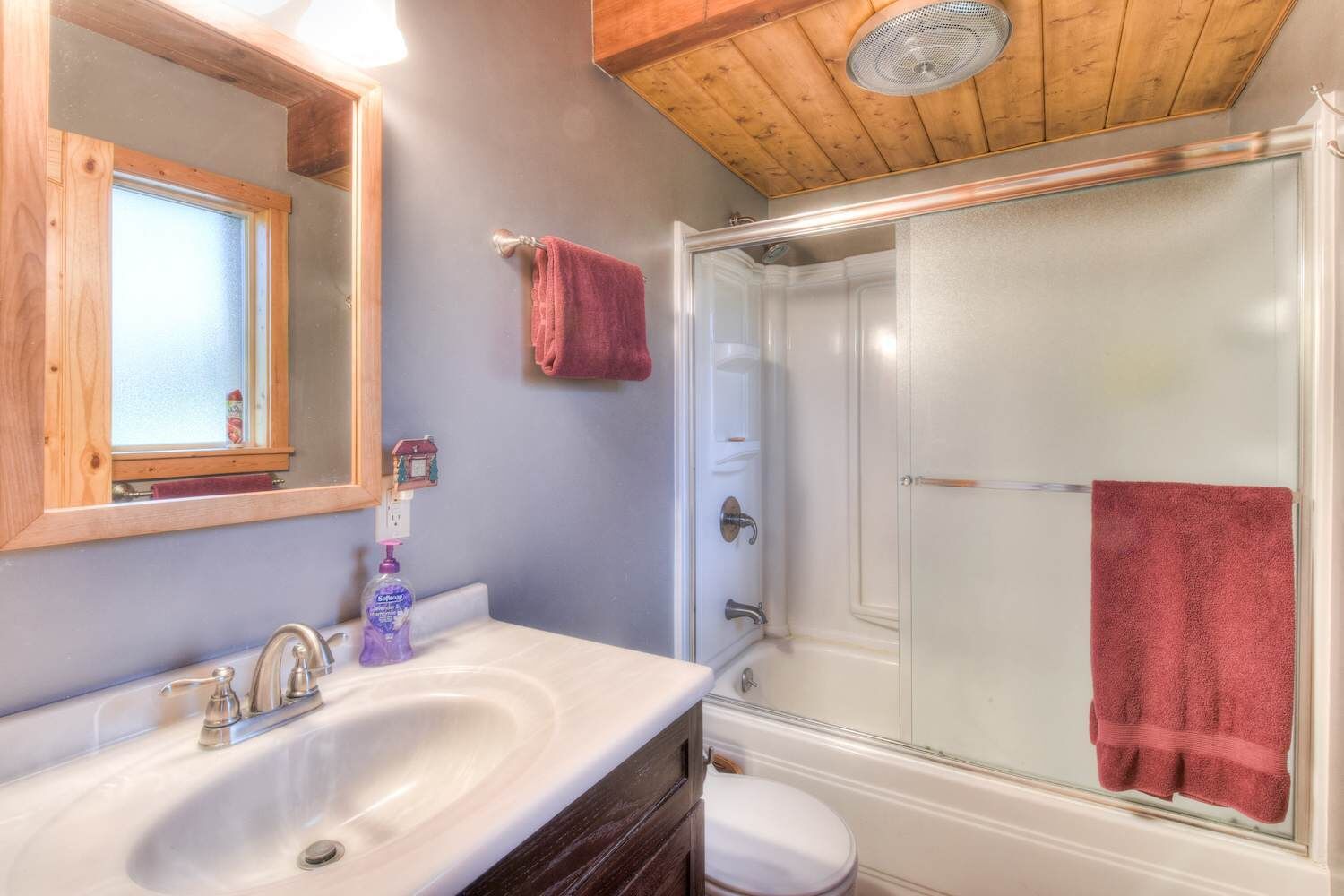 A bathroom with a wooden ceiling, a large mirror, a sink with a faucet, a toilet, and a glass-enclosed shower with a red towel hanging on the door.