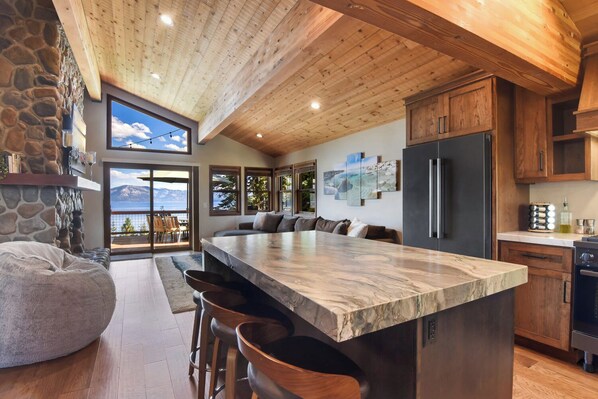 A modern kitchen with a large island, stone wall, wooden ceiling, and large windows overlooking a scenic view. - Shoreline Vista | 180° Lake Views | Pet-Friendly | 2 Master Suites (Carnelian Bay)