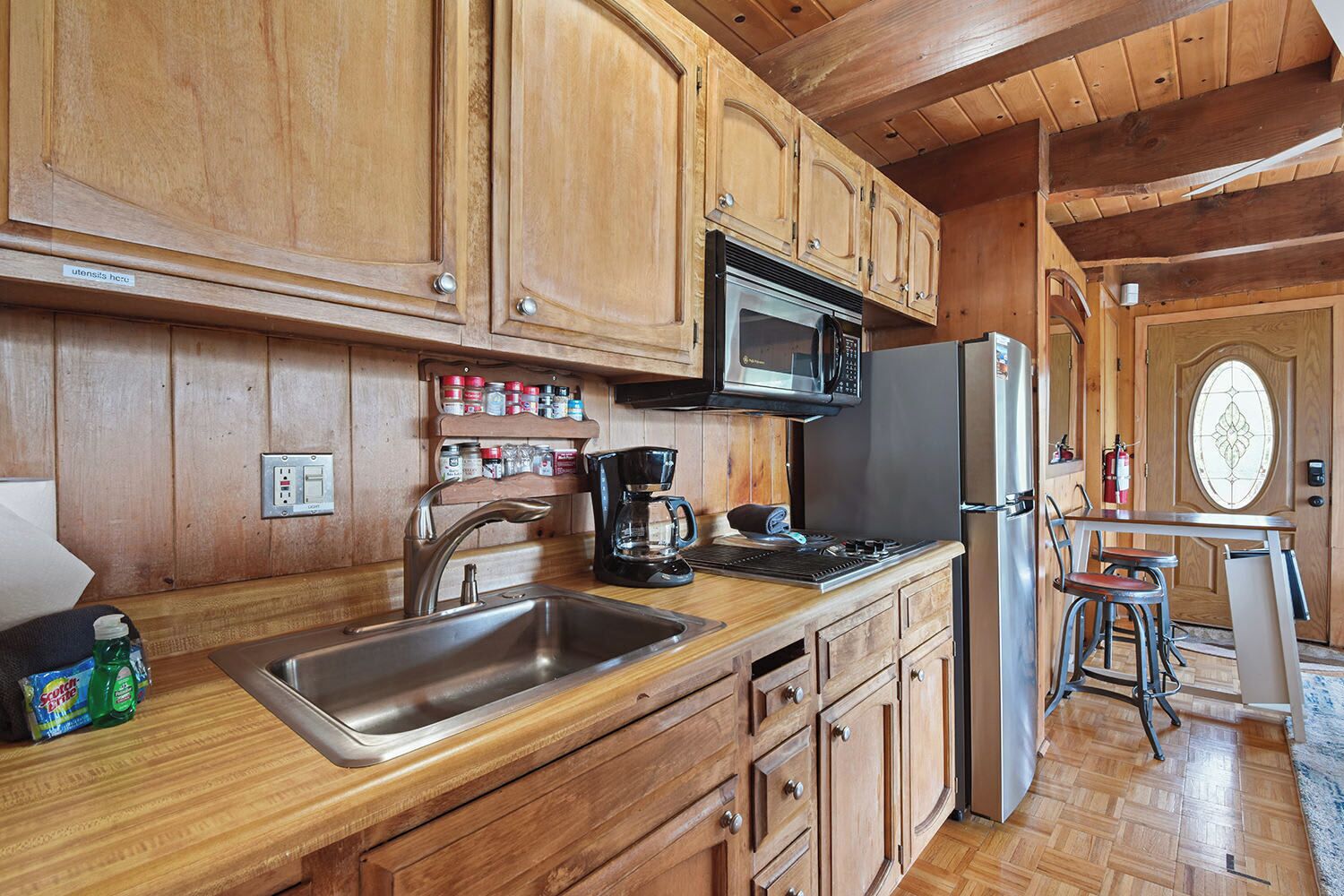 A wooden cabin kitchen with a stainless steel sink, a microwave, a stove, and a refrigerator. There is a shelf with spices and a coffee maker on the counter.
