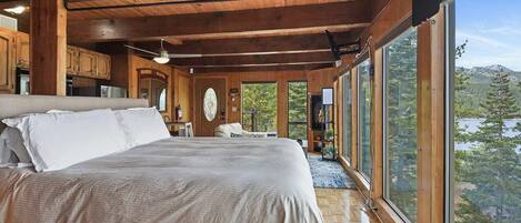 A spacious bedroom with a large bed, wooden ceiling beams, a fireplace, and a view of a forest and lake.