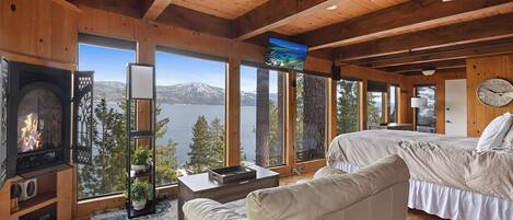 A cozy cabin room with a fireplace, a bed, a sofa, and large windows offering a view of a lake and mountains.