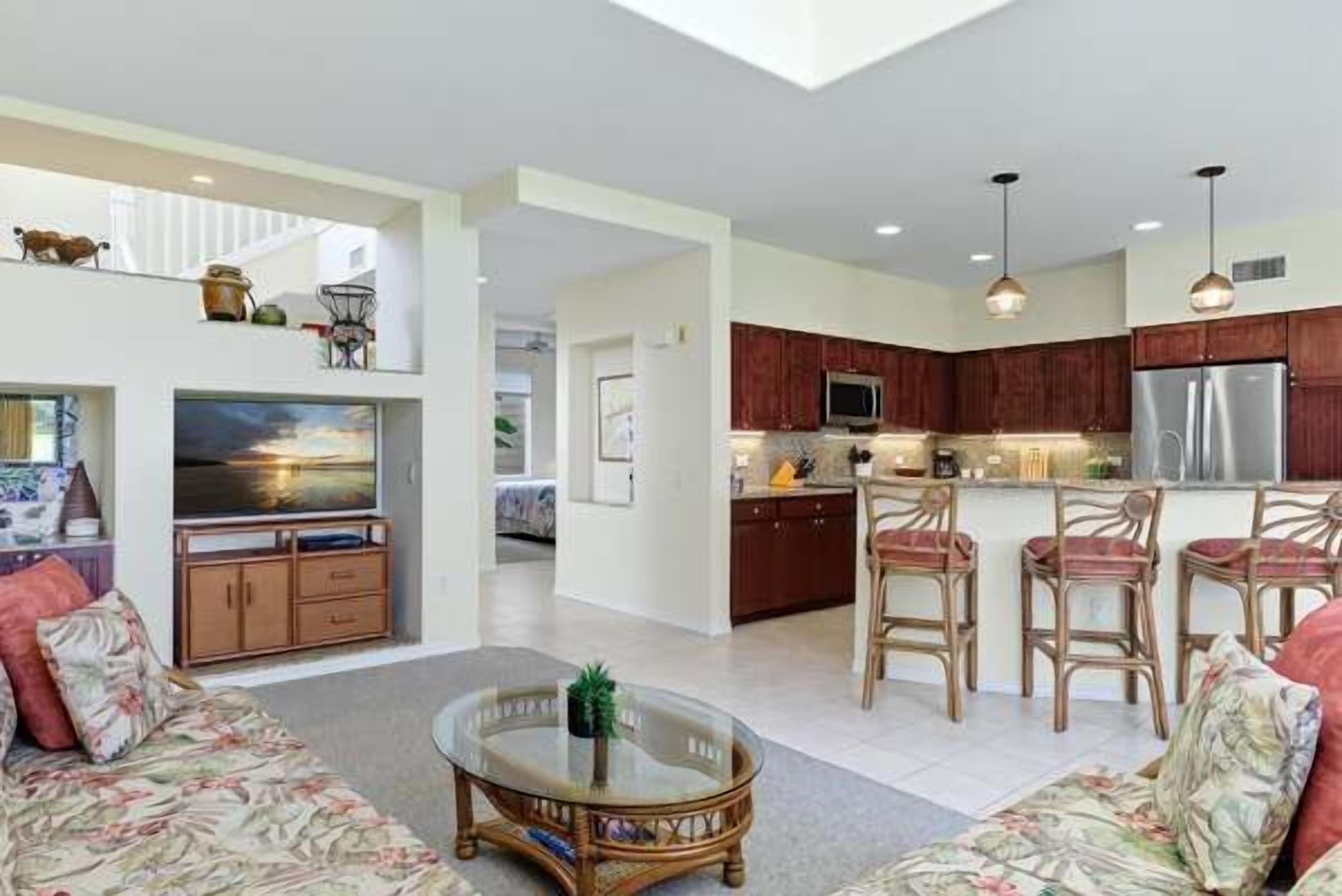 A spacious living area with a sofa, a glass coffee table, and a kitchen with wooden cabinets and a stainless steel refrigerator.
