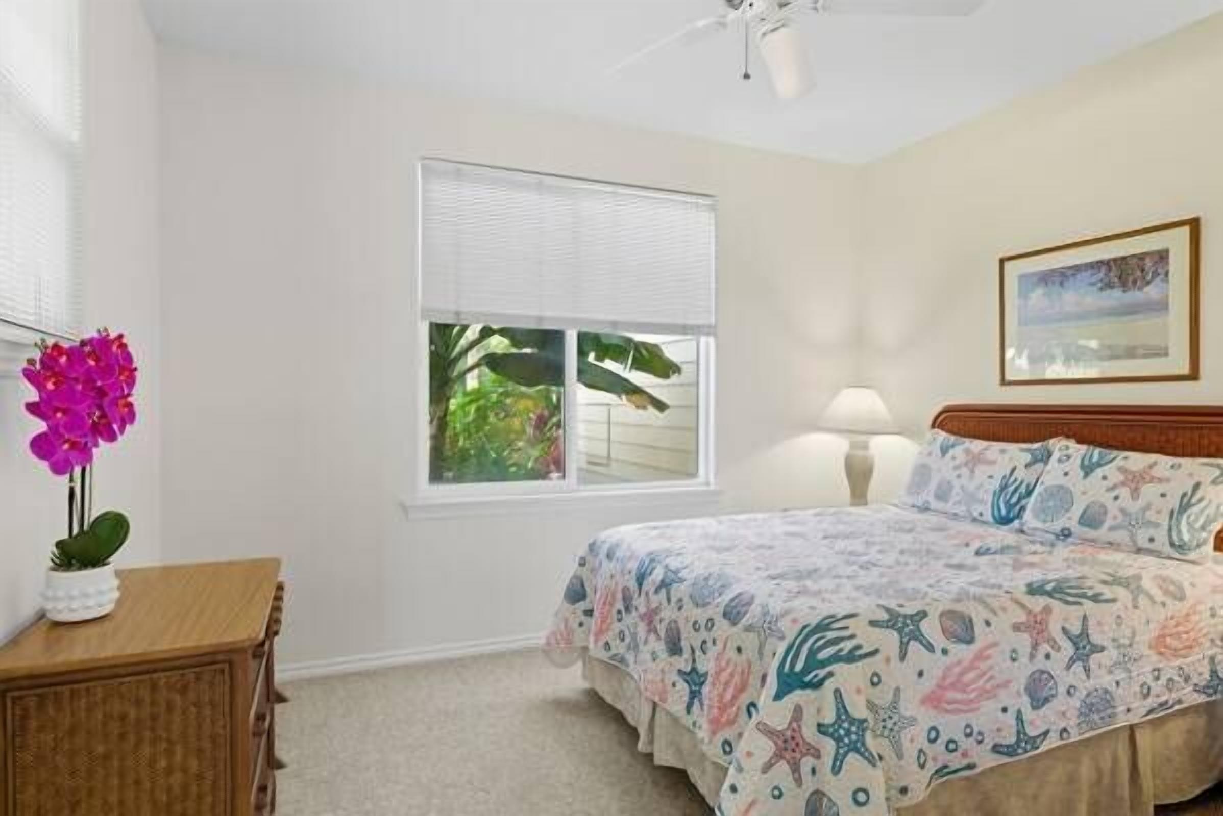 A bedroom with a bed, a bedside table, a lamp, a window with blinds, and a framed picture on the wall.