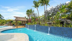 Ein Swimmingpool mit blau gefliestem Rand, einer Reetdachkonstruktion und üppiger tropischer Vegetation.