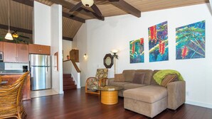 Ein modernes Wohnzimmer mit Sofa, Korbsessel, Glastisch und einer Küchenzeile inklusive Kühlschrank. Drei Bilder hängen an der Wand, und eine Treppe führt zu einer weiteren Ebene.