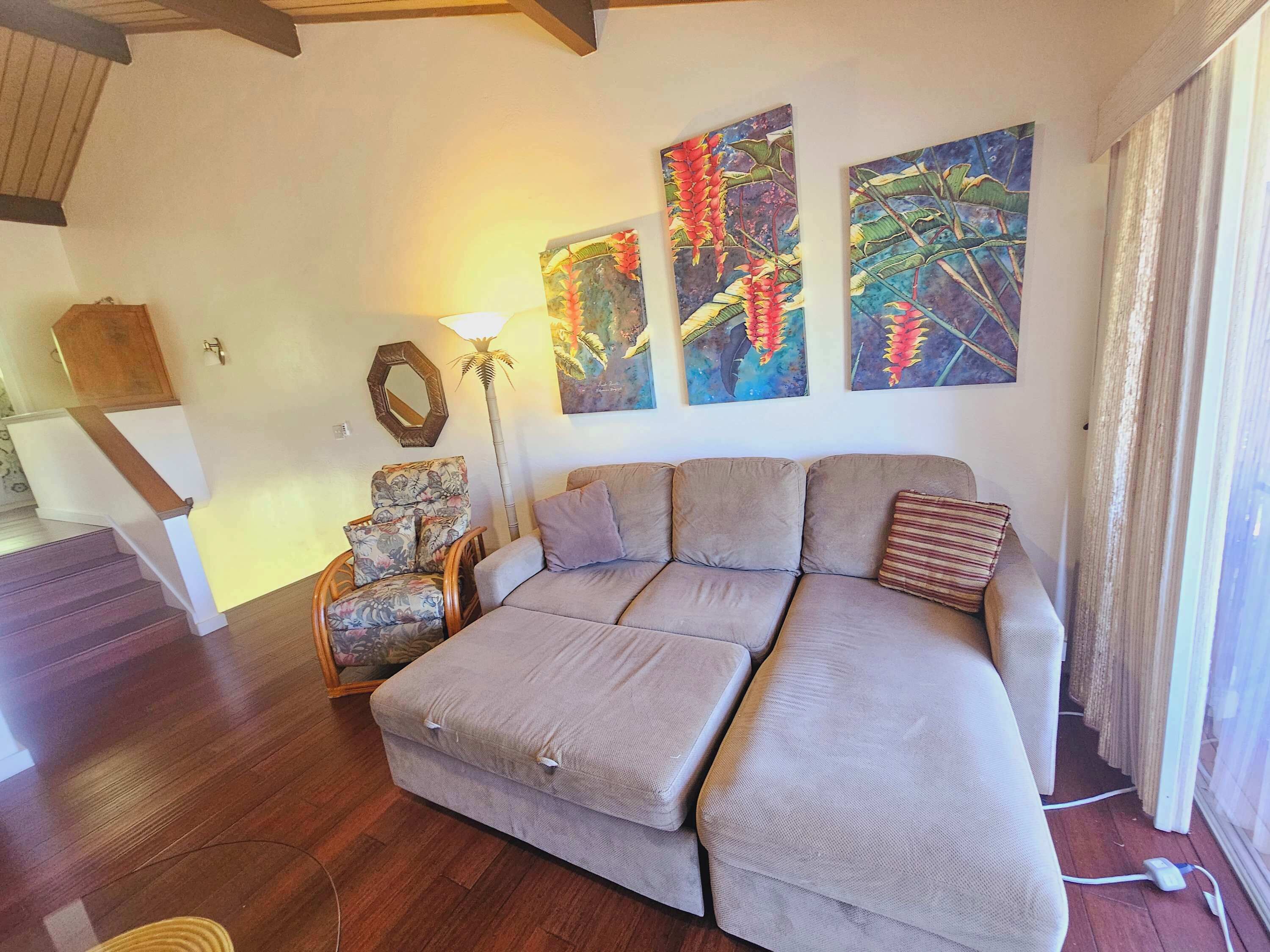 A living room with a beige sectional sofa, a wooden floor, a mirror, and three wall-mounted paintings of tropical flowers.