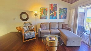 Ein Wohnzimmer mit einer Couch, einem Sessel und einem Glastisch. An den Wänden hängen drei Bilder und eine Stehlampe steht im Raum.