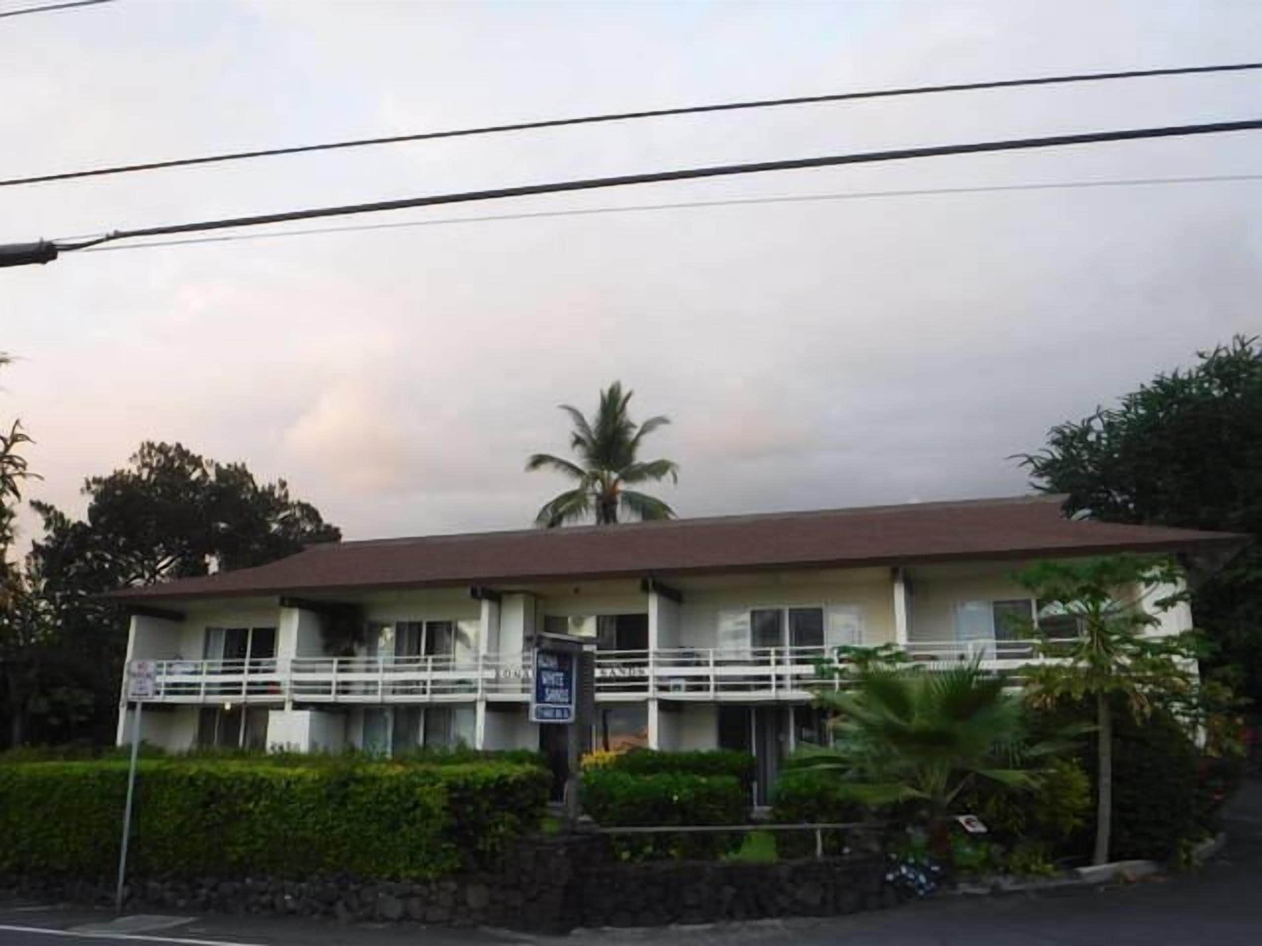A two-story building with a balcony on the upper floor, surrounded by greenery and palm trees.