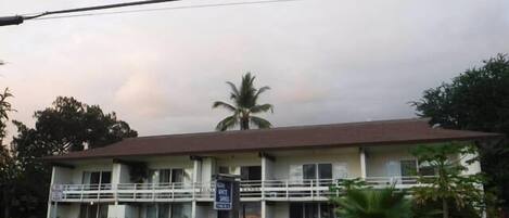 A two-story building with a balcony on the upper floor, surrounded by greenery and palm trees.