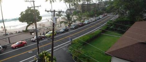 A street with cars parked on the side, a beach in the background, and a building with a brown roof.