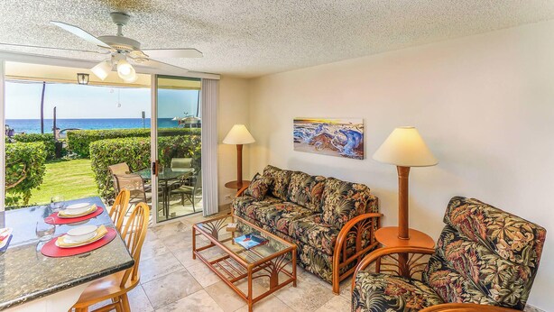 A living room with a sofa, chairs, a coffee table, and a dining table set for two. There is a ceiling fan, a painting on the wall, and a view of the ocean through large glass doors. - Kona White Sands A4 Romantic Ocean View! (Kailua-Kona)
