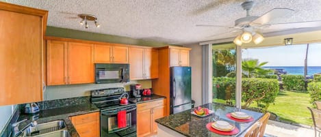A kitchen with wooden cabinets, a microwave, an oven, and a refrigerator. There is a dining area with a table and chairs, and a view of the ocean through large windows.