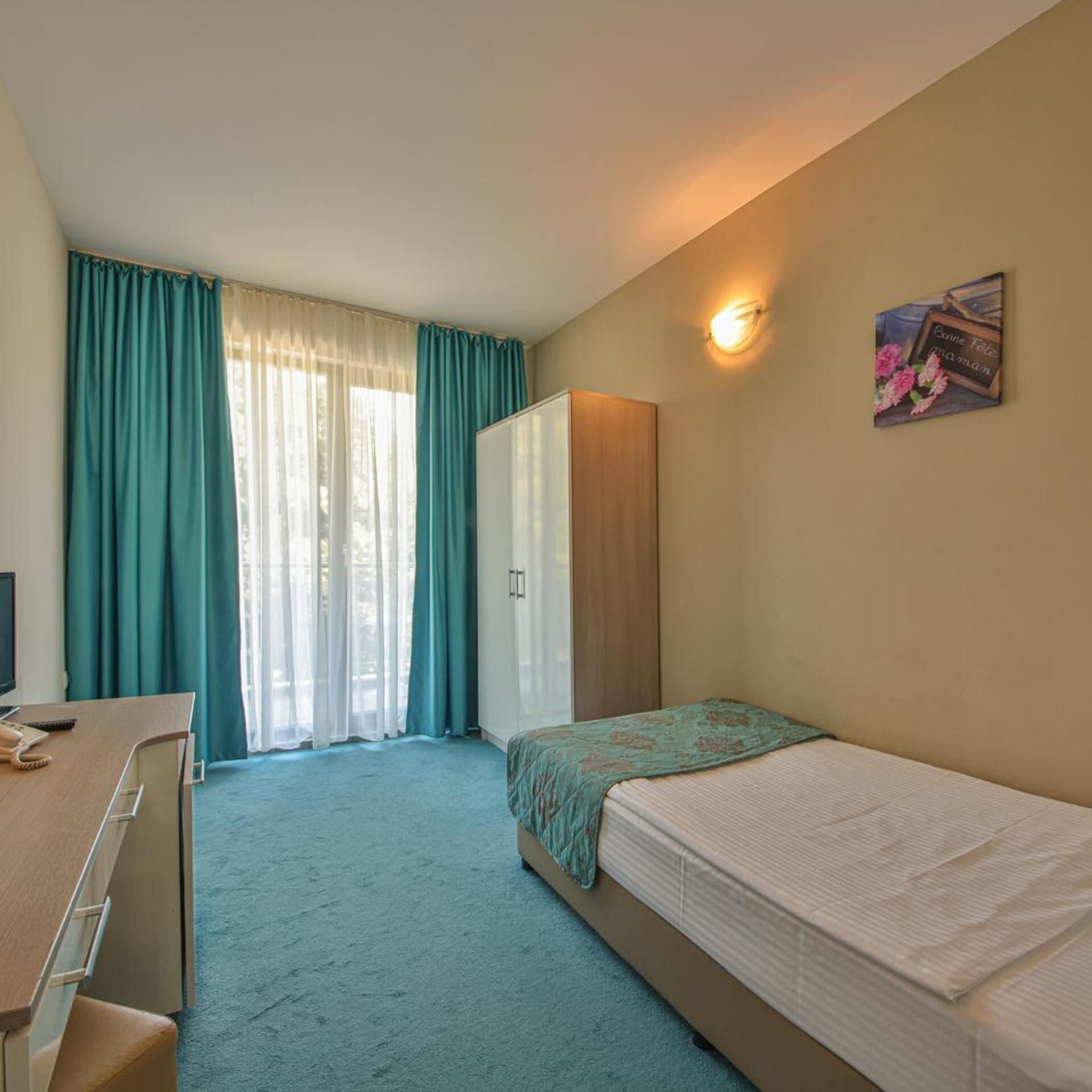 Quarto de hotel com cama, escrivaninha, guarda-roupa e janela com cortinas.