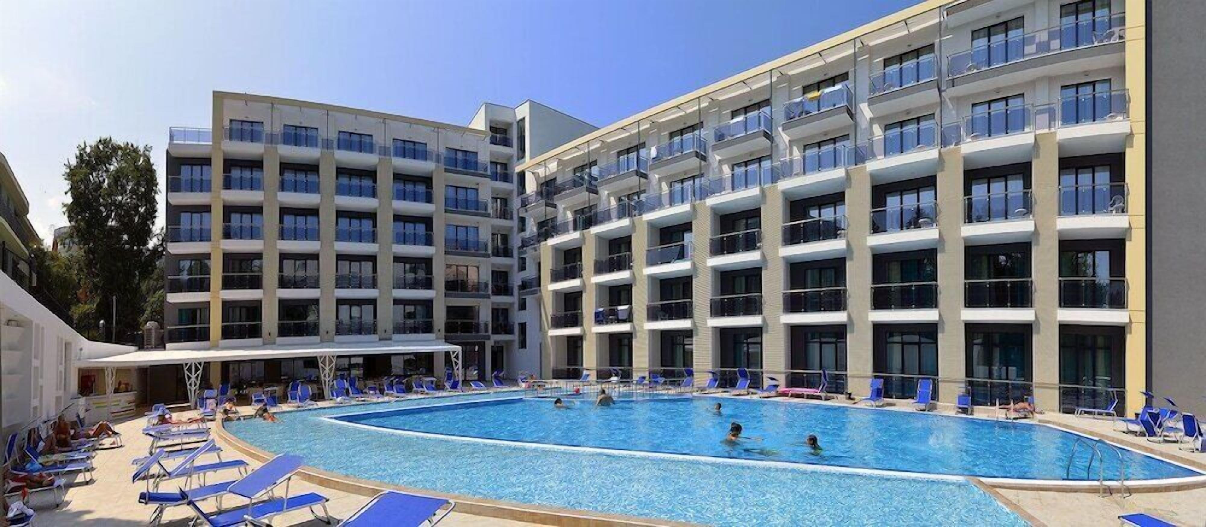 A hotel with a large swimming pool surrounded by sun loungers and a multi-story building with balconies.