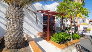 Un edificio blanco con puerta y ventanas rojas, una palmera y un pequeño jardín con plantas.