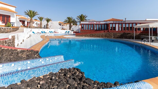 Una piscina rodeada de edificios blancos, un bar junto a la piscina y una pizzería.