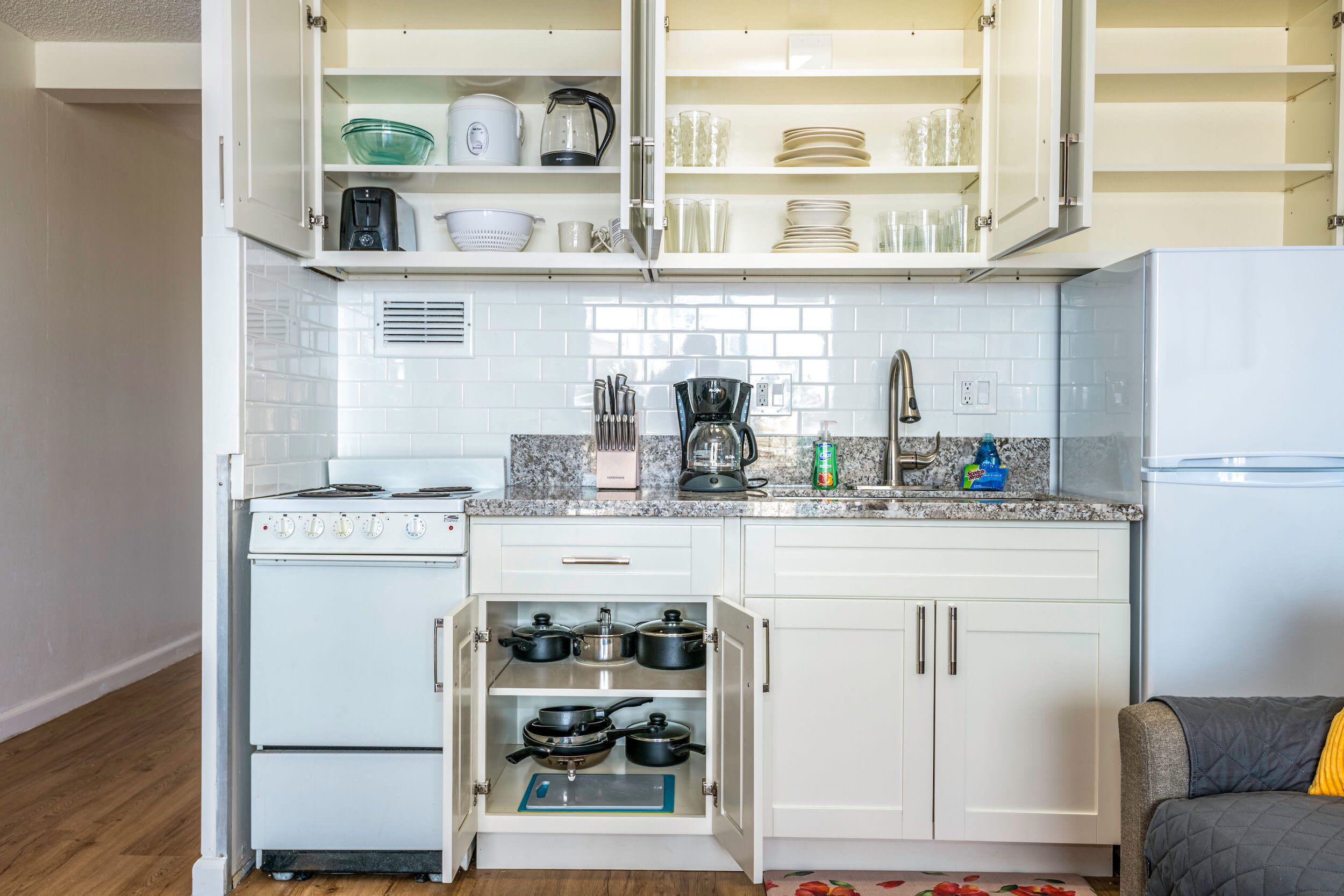 A compact kitchen with white cabinets, a white stove, and a built-in oven. There is a countertop with a coffee maker, a kettle, and a sink.