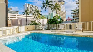 A clear blue pool with lounge chairs and palm trees, surrounded by modern buildings.
