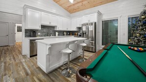 Een moderne keuken met witte kasten, een centraal eiland met barstoelen, een roestvrijstalen koelkast en een pooltafel.