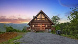 A log cabin with a porch, a gravel driveway, and a mountainous backdrop. - Sky Views - 2BR/New Hot Tub/New Firepit/Grill/Sleeps 8 (Gatlinburg)