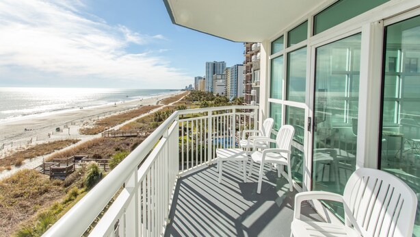 Un balcone con sedie bianche che si affaccia su una spiaggia e l'oceano, con edifici alti sullo sfondo.