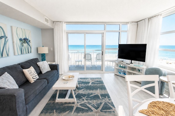 A living room with a sofa, coffee table, TV, and a view of the beach through large windows.