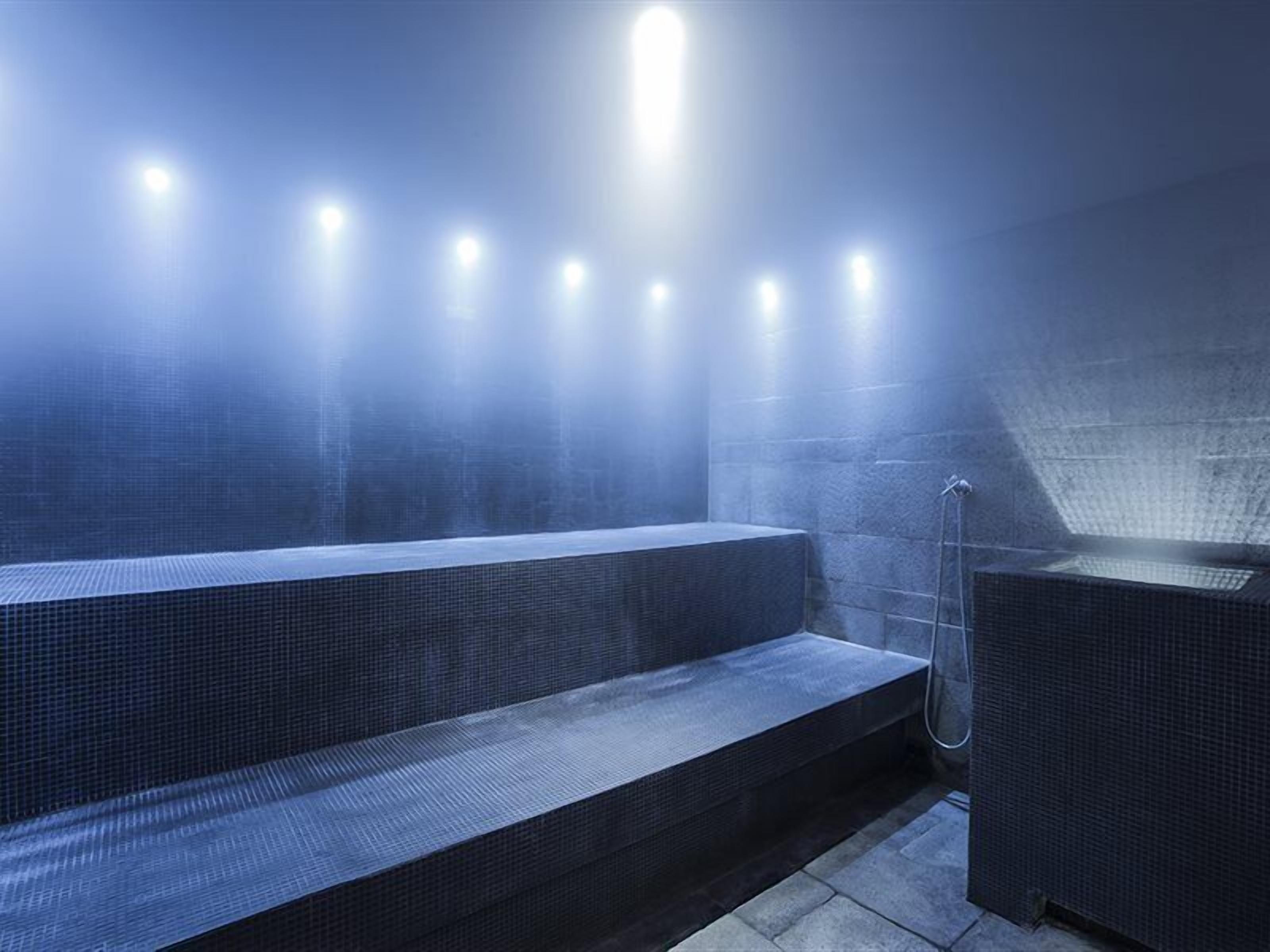 Uma sauna moderna, com iluminação suave, possui um banco e um espaço separado para vapor, com piso ladrilhado e parede de pedra.