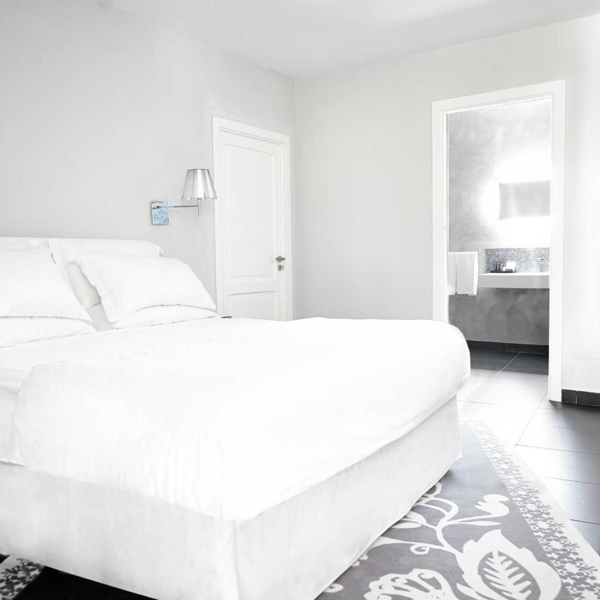 A bedroom with a bed, a door, a nightstand with a lamp, and a bathroom visible through an open door.