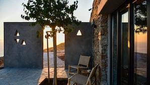 A stone patio with two wicker chairs, a tree, and a stone wall with triangular cutouts. - Under the Sun Cycladic Village (Tinos)