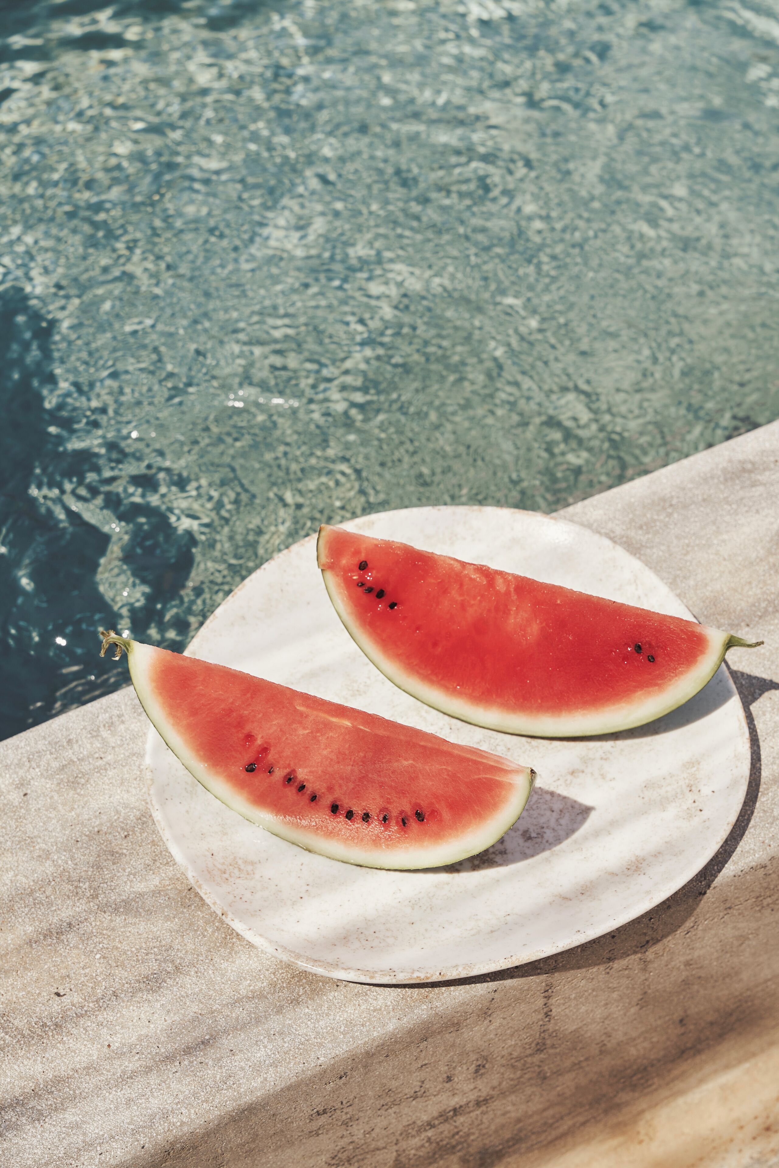 Two halves of a watermelon on a white plate by a pool.