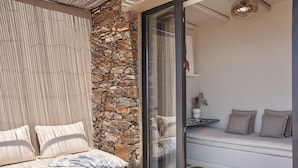 A cozy seating area with a stone wall, a cushioned bench, and a glass door leading to an outdoor space. - Under the Sun Cycladic Village (Tinos)