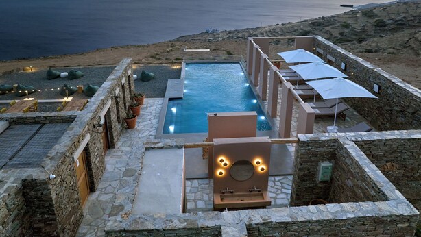 A pool area with stone walls, lounge chairs, and umbrellas, overlooking a body of water. - Under the Sun Cycladic Village (Tinos)