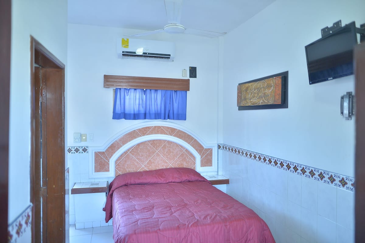 A bedroom with a bed, an air conditioner, a ceiling fan, a window with curtains, and a wall-mounted TV.