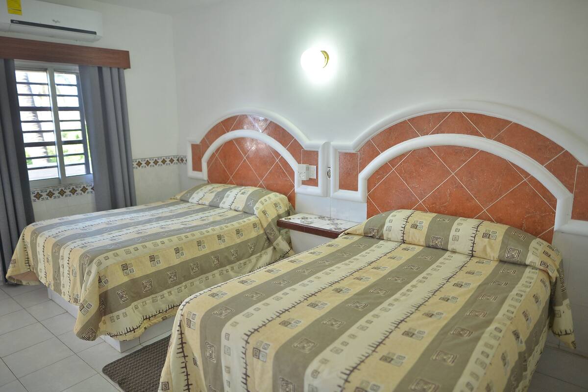 A hotel room with two beds, each with patterned bedding, a nightstand, and a wall-mounted light fixture.