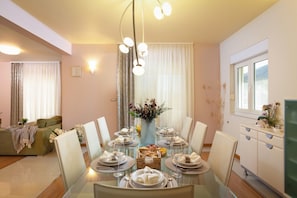 A dining area with a glass table set for six, surrounded by white chairs, with a sofa and a cabinet in the background. - Luxury Villa Lovric Private Heated Pool (Omis)