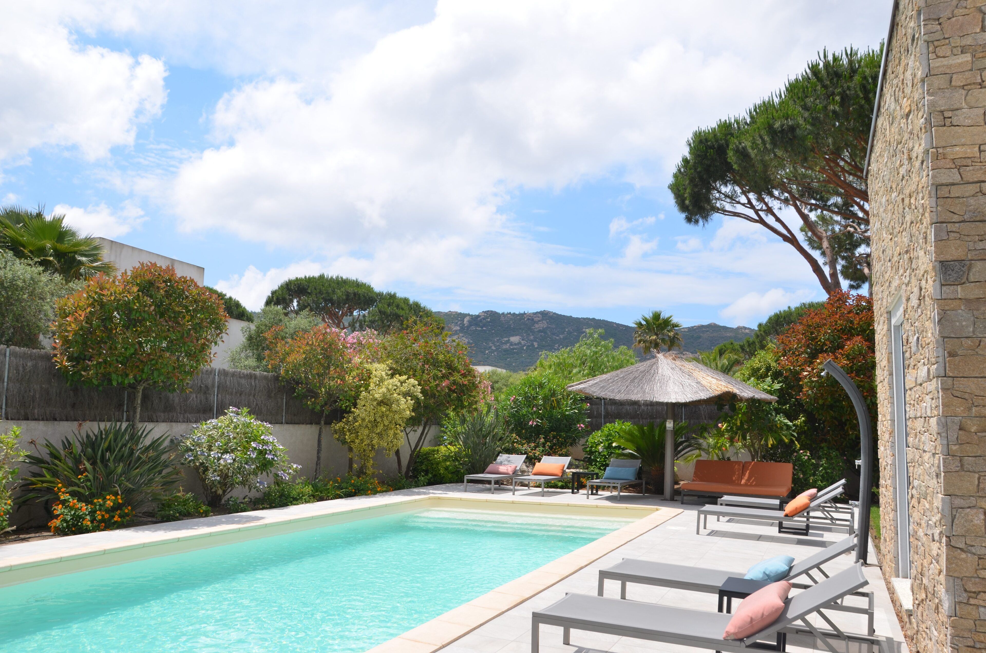 A swimming pool with lounge chairs and a thatched umbrella, surrounded by trees and a stone building.