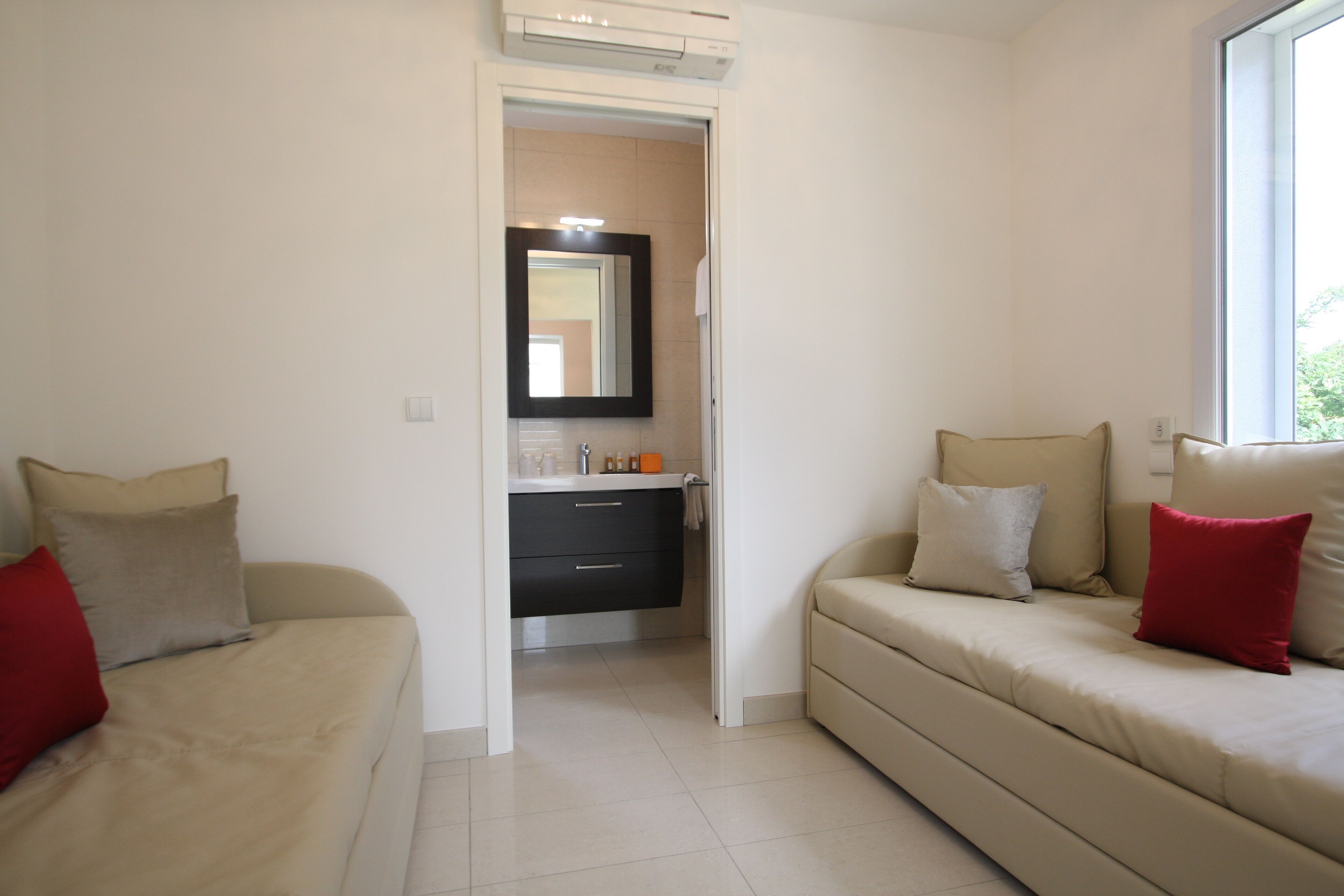 A room with two sofas, one with red and beige cushions, and another with beige and red cushions. There is a small bathroom with a mirror and a sink.
