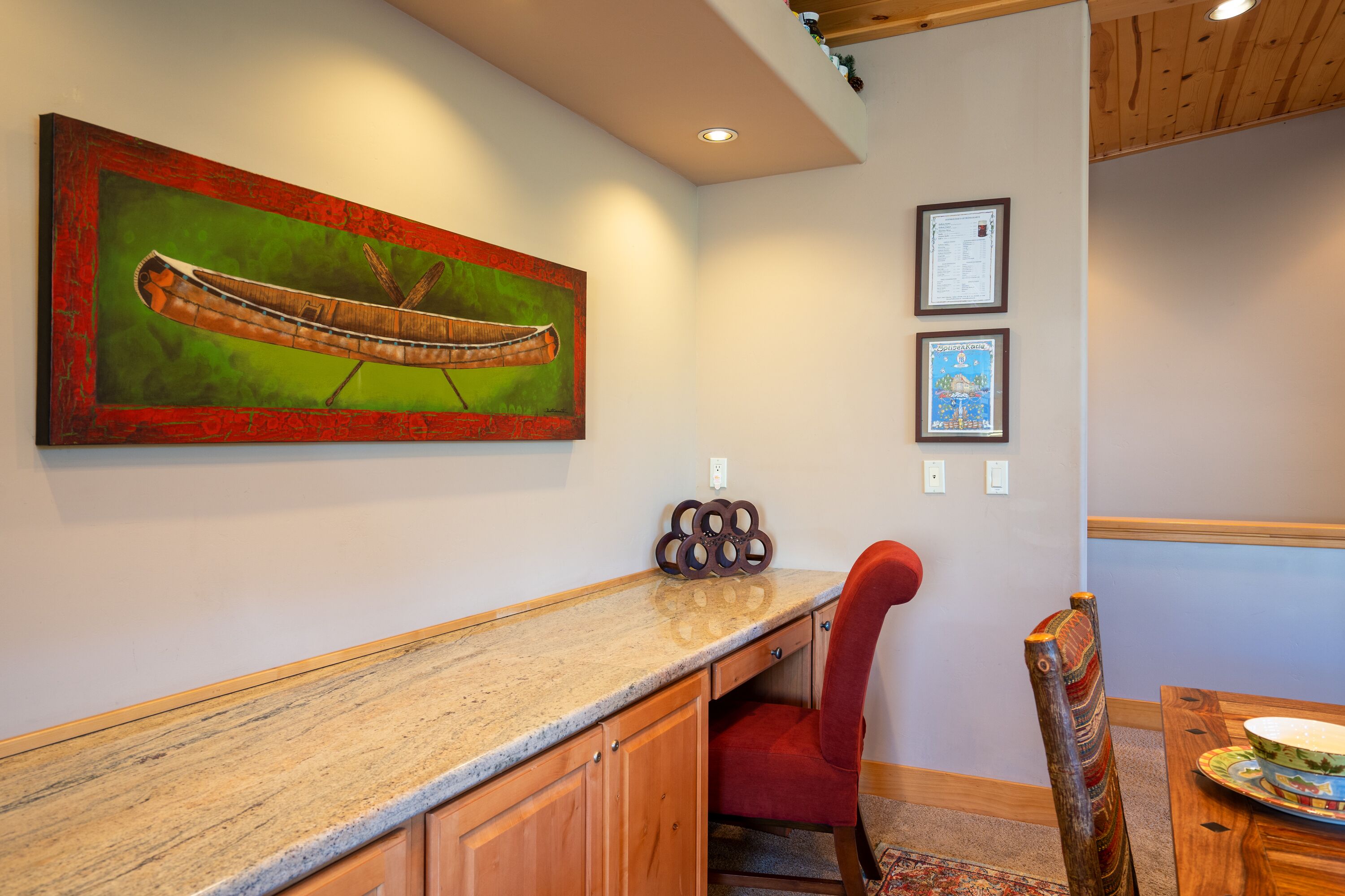 A dining area with a wooden table, a red upholstered chair, a granite countertop, and a canoe painting on the wall.
