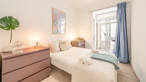Ein Schlafzimmer mit Bett, Nachttischen, einem Schminktisch und einem großen Fenster mit Vorhängen sowie Blick ins Freie.