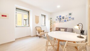Ein geräumiges Zimmer mit einem runden Esstisch, einer gemütlichen Couch und einem Sessel. Zwei Fenster mit Vorhängen, ein Wandbehang aus Makramee und ein Regal mit Dekorationsobjekten.