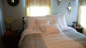 A neatly made bed with white linens and pillows, a bedside table with a clock and a book, and a wall-mounted candle holder. - Historic Century Home on Chardon Square (Chardon)