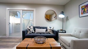 A living room with a dark blue sofa, a white armchair, a wooden coffee table with a decorative bowl, a floor lamp, and a large mirror on the wall. - Cozy Remodeled Apt Home w/Patio & Great Location (Austin)