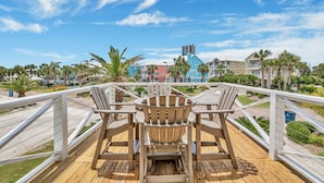 Um deck de madeira com cadeiras e uma mesa, com vista para uma praia com palmeiras e edifícios coloridos.
