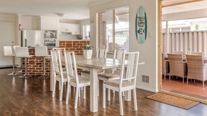 Un coin repas avec une table en bois et des chaises blanches, une cuisine en arrière-plan et une vue à travers une porte.