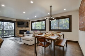 Uma sala de estar moderna com lareira, canto de jantar com mesa e cadeiras de madeira, e janelas amplas com vista para árvores.