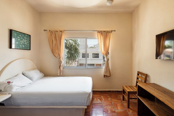 A bedroom with a bed, a wooden bench, a window with curtains, a wall-mounted TV, and a framed picture.